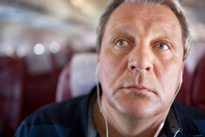A male passenger with earphones looks forward on an airplane, potentially a passenger doing things that make flight attendants roll their eyes.