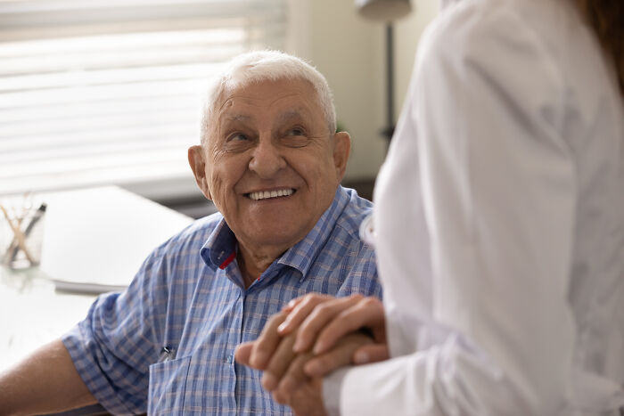 An older man smiles brightly at a doctor, who holds his hand, illustrating various pain reactions in patients.