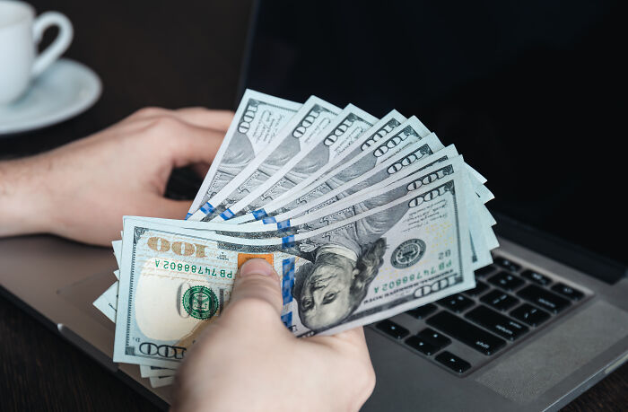 A hand holding US dollar bills in front of a laptop, highlighting marriage regrets