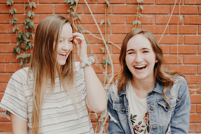 Two young women laughing together against a brick wall, symbolizing a fake friend dynamic