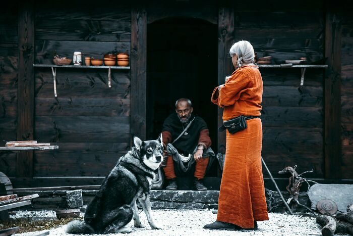 A woman in historical costume with a dog in front of a wooden house, depicting historical facts.