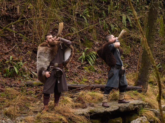 Two men in historical Viking-like attire blowing horns in a forest, related to historical facts.