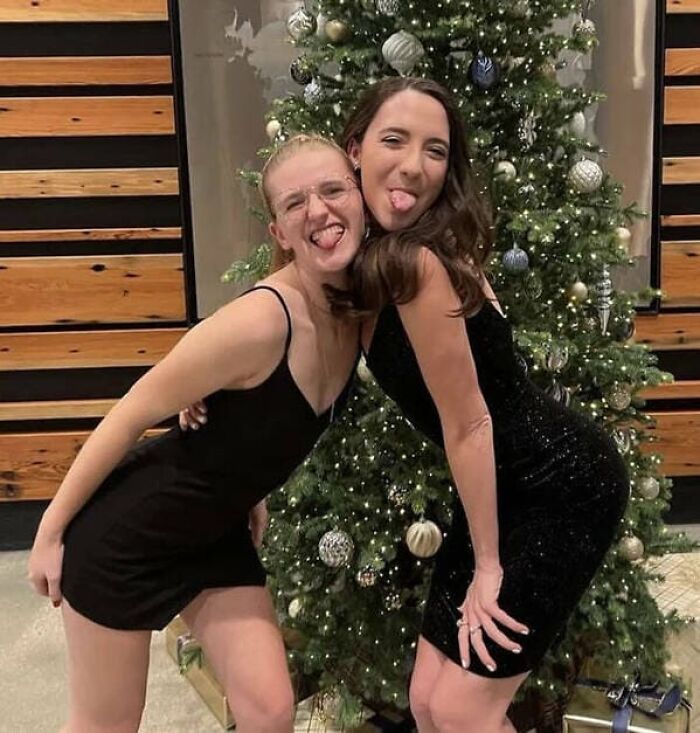 Two young women posing playfully in black dresses next to a Christmas tree, making you look twice.