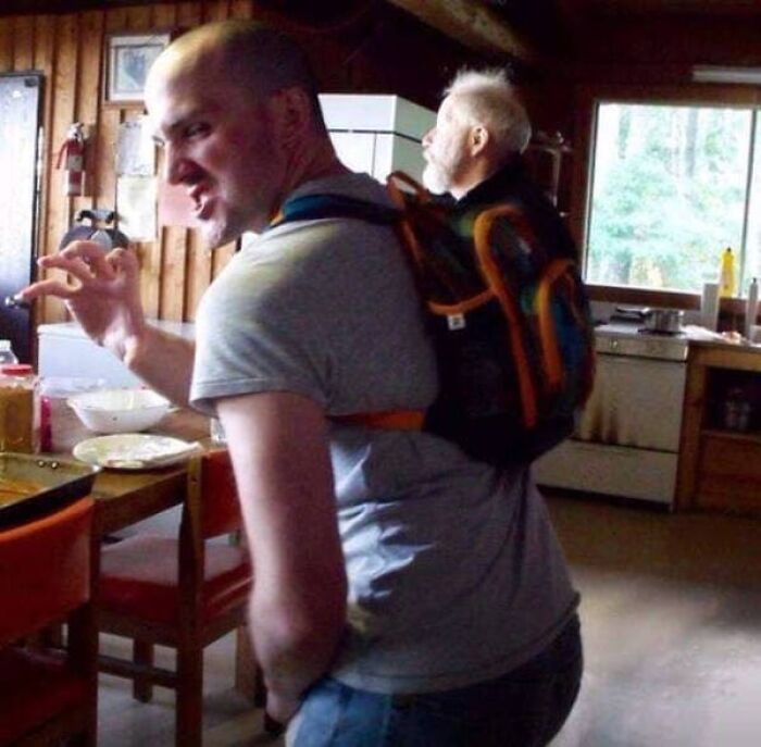 A confusing picture where an older man looks like he is riding inside the backpack of a younger man.