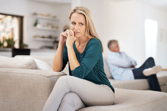 Woman looking worried on a couch, man in background, reflecting on marriage and divorce.