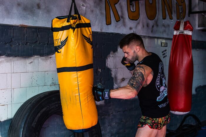 A male coworker in a black tank top and boxing gloves practicing punches on a yellow heavy bag