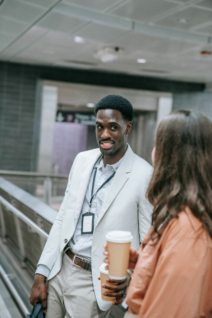A male coworker in a light suit holding a coffee cup and speaking to a female colleague