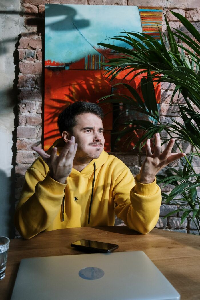 A male coworker in a yellow hoodie looking confused while shrugging with hands up in a cafe