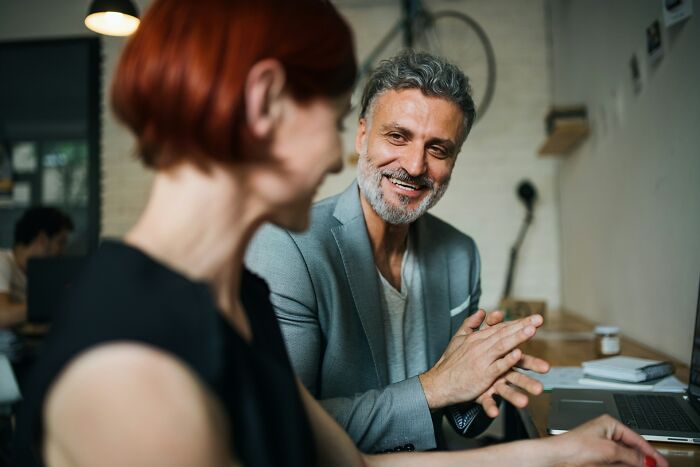 A senior male coworker smiling while talking to a female colleague in an open office space