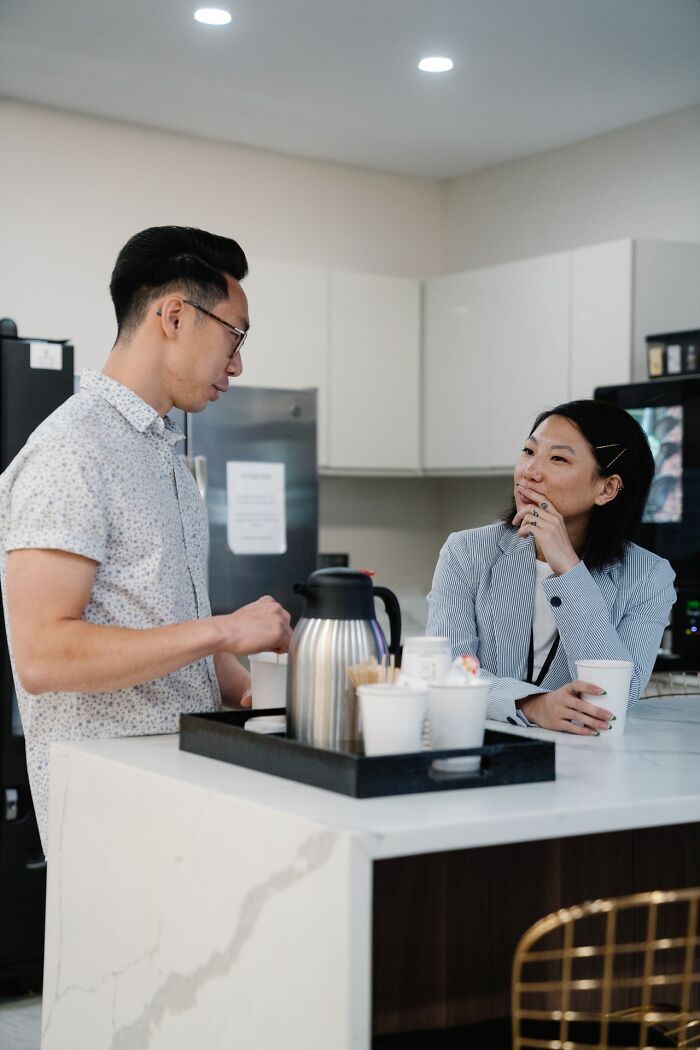 An Asian female coworker listening to a male colleague in an office kitchen during a break