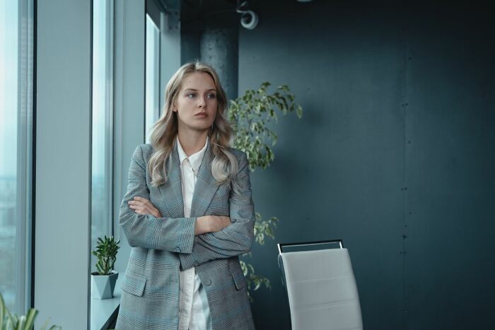A thoughtful female coworker standing near an office window with arms crossed in a modern workplace setting
