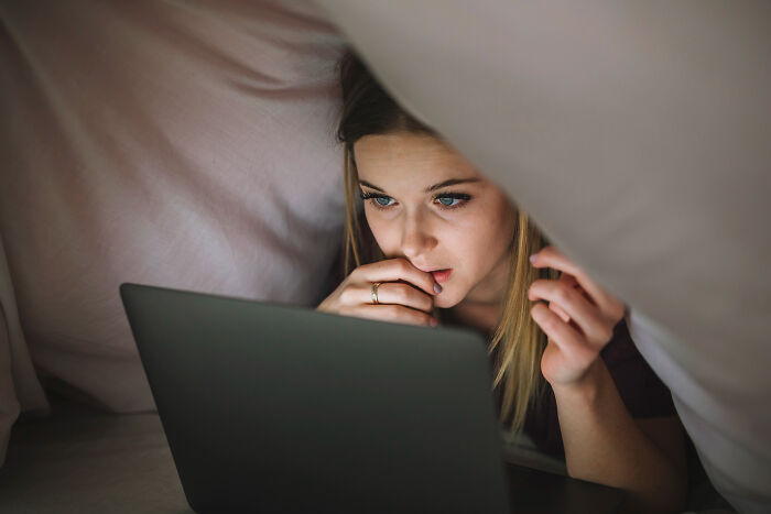 A worried woman secretly using a laptop under a blanket, representing a creepy secret regarding a partner.