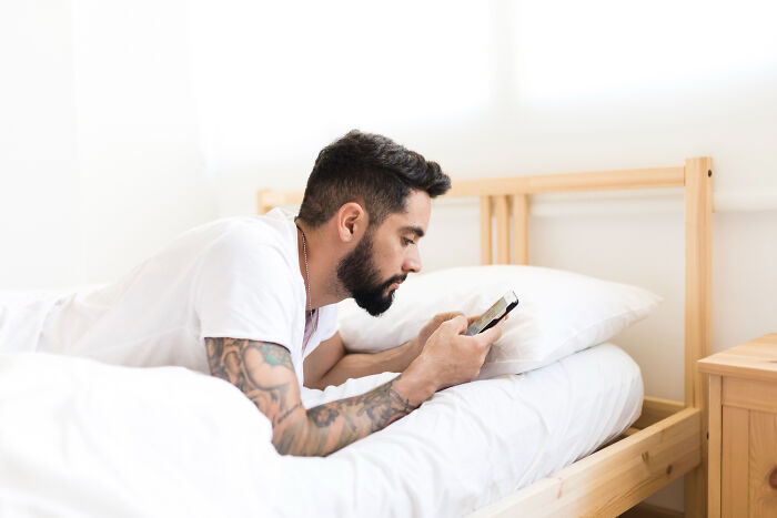 A bearded man with tattoos lying in bed, looking at his phone to hide creepy things done to a partner