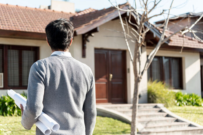 A man holding blueprints looks at a house, thinking about things to do to a partner's home.