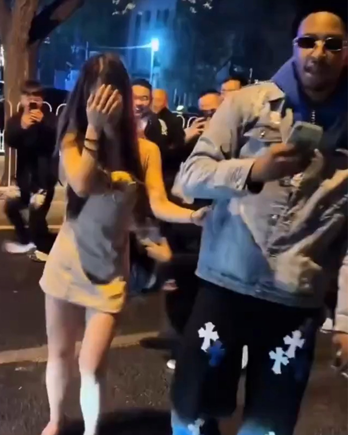 Woman covering her face in public, surrounded by a crowd as a shaming trend spreads for dating a foreigner.