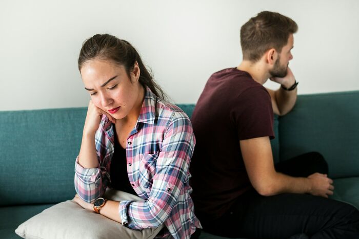 A sad young couple sitting back to back on a couch, highlighting romance and relationship challenges.