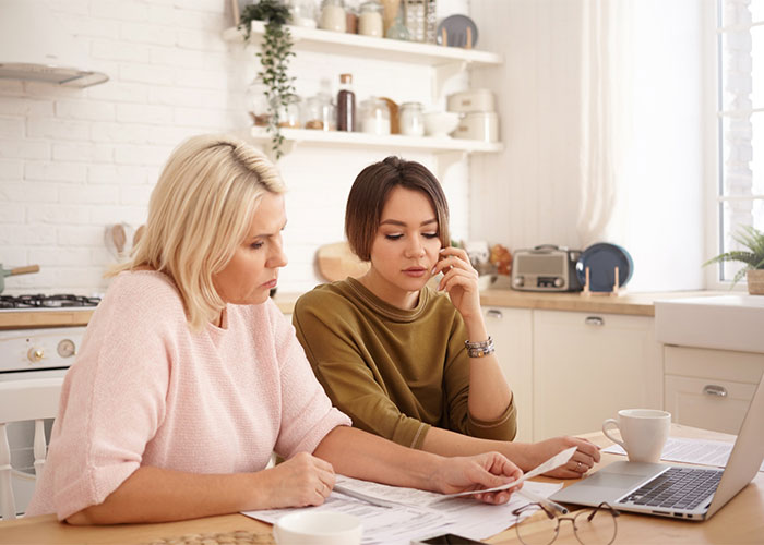 Two women look at documents with a laptop, reflecting the stress of wedding drama and gift cards.