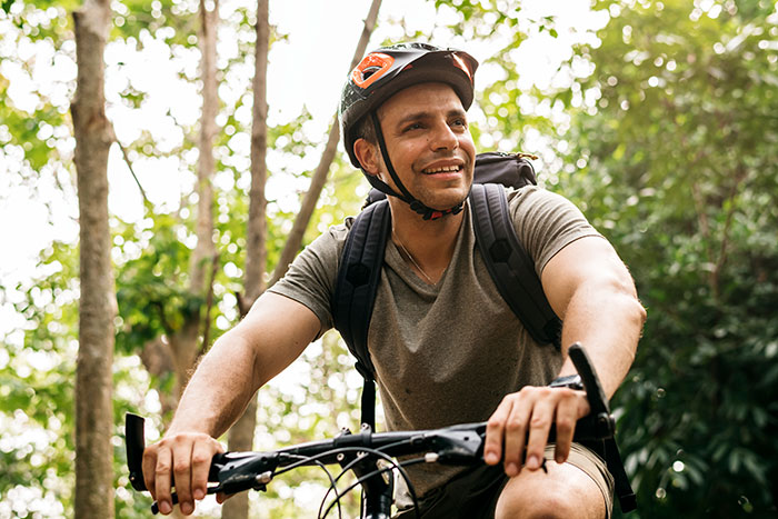 A man in a helmet smiles while biking through a forest, experiencing wild delayed plot twists in his life.