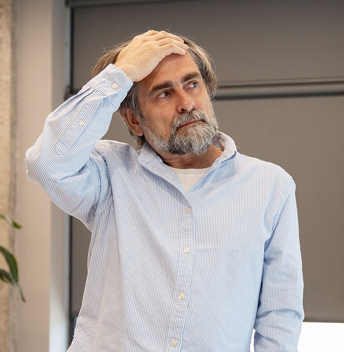 A man with a beard and a light blue striped shirt, holding his head, affected by a wild delayed plot twist.