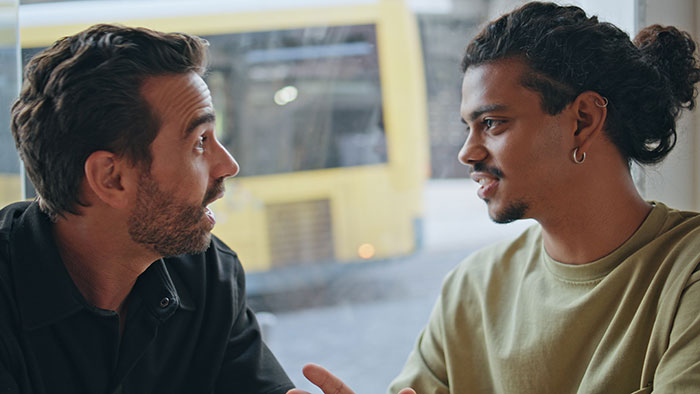 Two men in profile are deep in conversation by a window, discussing wild plot twists from their lives.