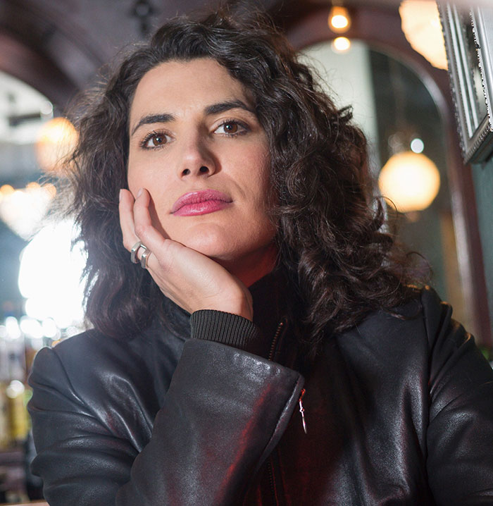 A woman with dark curly hair and a black leather jacket, contemplating a delayed plot twist from her life.