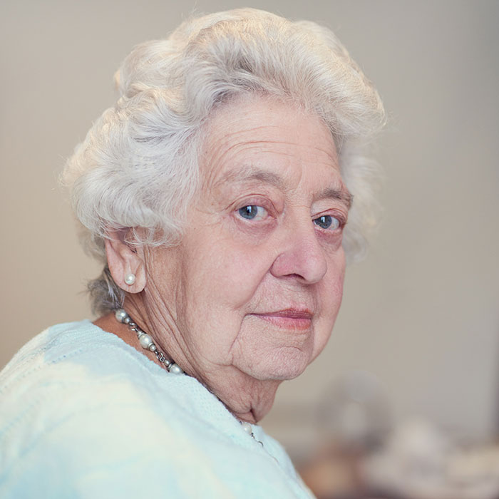 An elderly woman with white curly hair and pearl earrings, experiencing a delayed plot twist in her life.