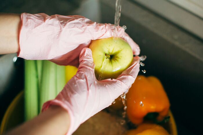 Hands in pink gloves washing a green apple under running water, highlighting the cleanliness expected, versus dirty truths industry insiders know.
