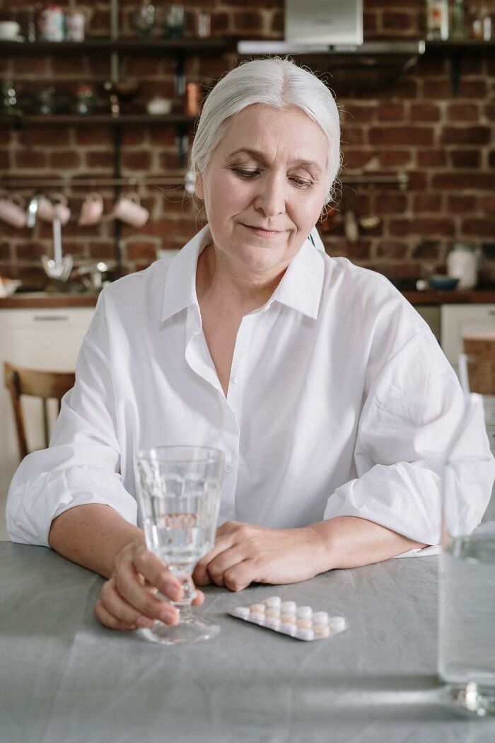 An older woman looks at pills on a table with a glass of water, reflecting on medical education choices.