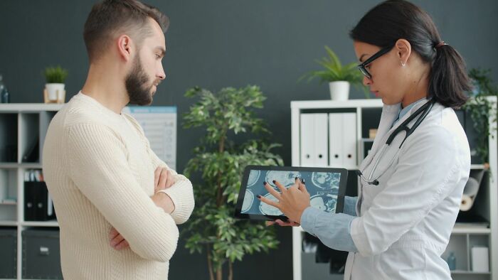 A male patient with arms crossed listening to a female doctor explaining medical results on a tablet.