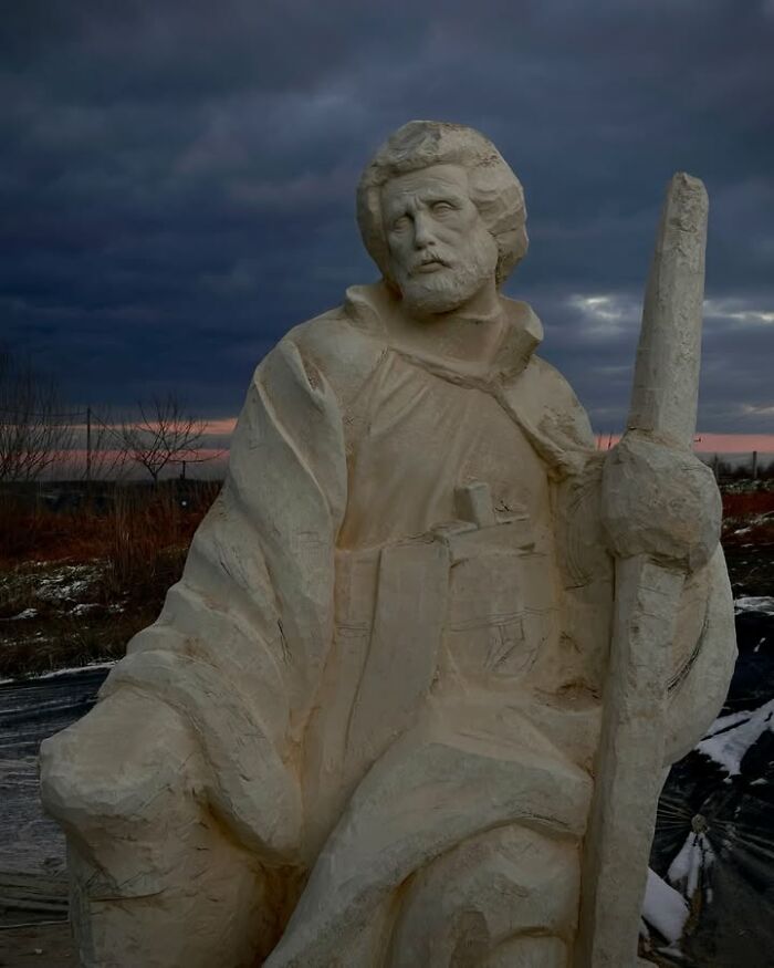 A large lifelike wood sculpture of a bearded saintly figure standing outdoors.