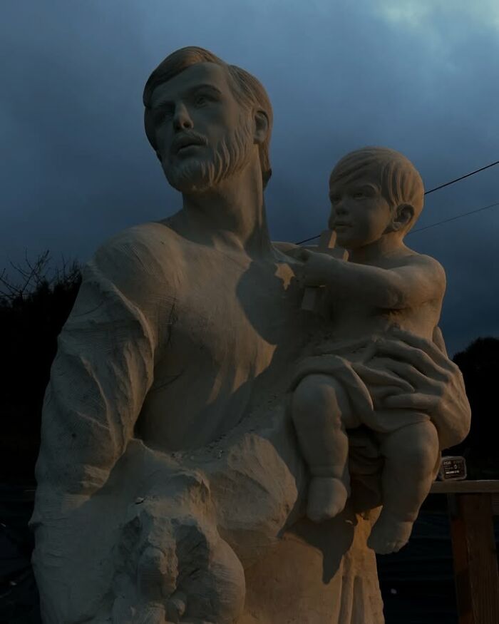 A Polish artist’s wood sculpture of a bearded man holding a child under a cloudy sky.