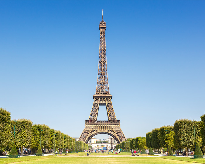 The Eiffel Tower in Paris, France, with green lawns and trees in the foreground under a clear blue sky, closed amid outrage.