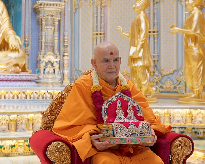 A Hindu guru, wearing orange robes and a mala, sitting on a red ornate chair, holding a deity, amid outrage.