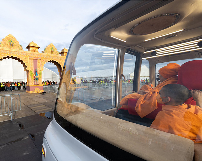 Hindu guru in orange robes in a vehicle, with an ornate temple gate and crowd visible outside, amid outrage.