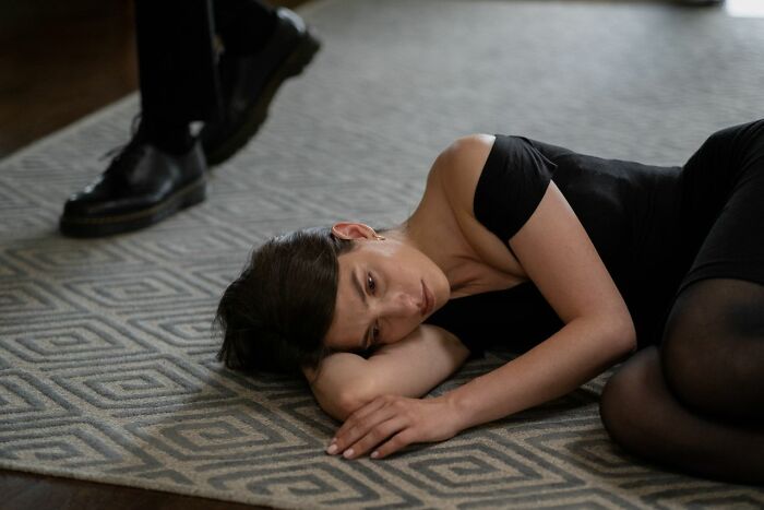A woman lies on a patterned rug, looking sad and defeated, while a person walks past her in the background, symbolizing the end of a marriage.