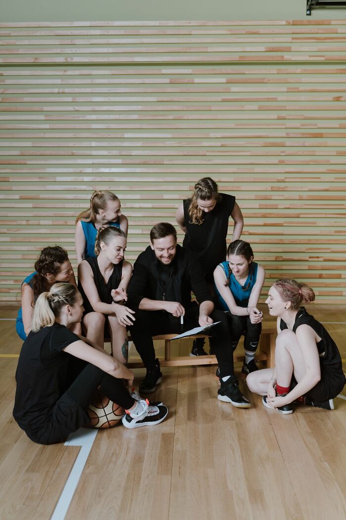 A group of women in athletic wear huddle around a male coach who is pointing at a clipboard, looking like a team that's getting a pep talk, illustrating marriage.