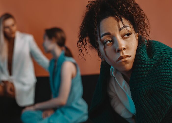 A woman with curly hair looking away sadly, while two blurred figures talk in the background, realizing friends were haters.