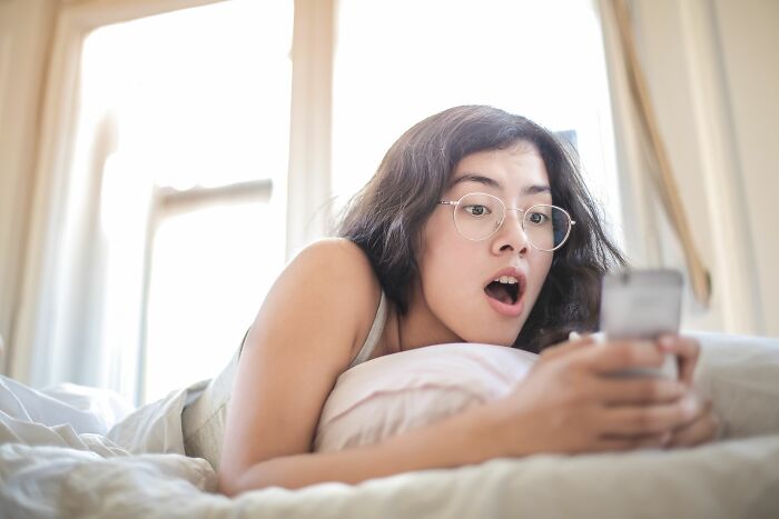 A surprised woman in bed looks at her phone, as if realizing friends were haters.