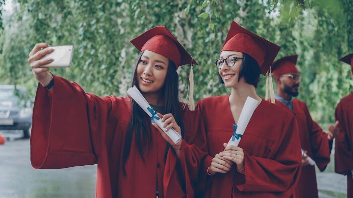 Two smiling graduates in red caps and gowns taking a selfie; a reminder that friends can be haters.