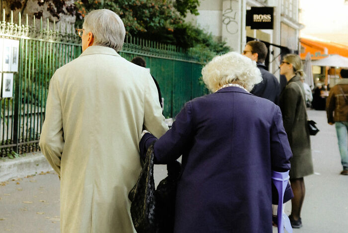 An elderly couple uncovers secrets while walking, the woman using crutches, as they walk away from the camera on a city street.