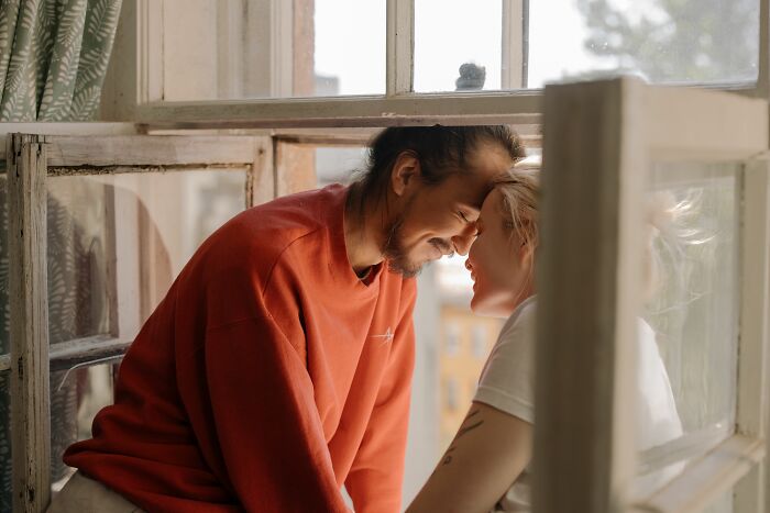 A loving couple uncovers secrets, facing each other through a window frame, their faces close and smiling.
