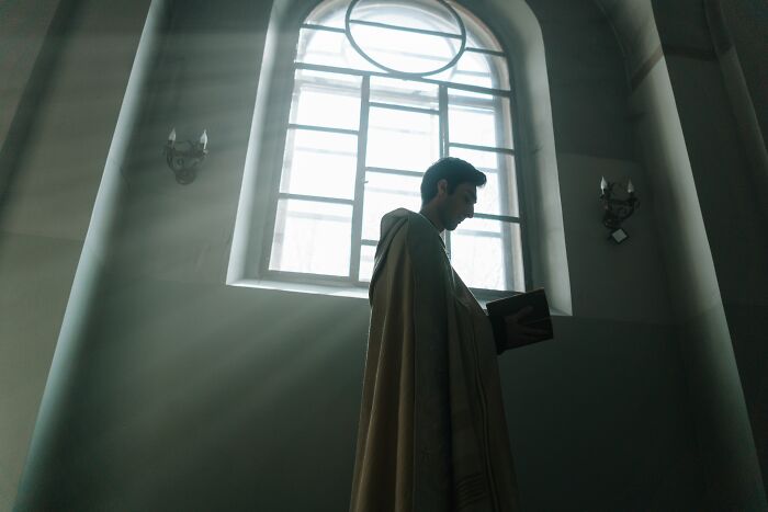A man in a cassock uncovers secrets, reading a book in a sunlit room, with light streaming through a tall window.