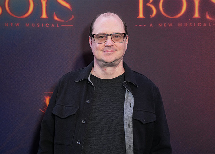 A man with glasses and a dark jacket smiles, standing against a backdrop for a musical production, related to Stephen King adaptation. A man with glasses and a dark jacket smiles, standing against a backdrop for a musical production, related to Stephen King adaptation.