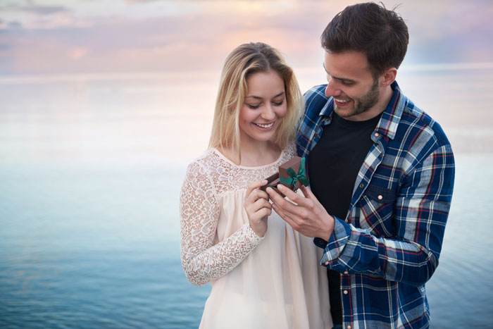 Couple smiling as man gives woman a ring outdoors by water at sunset Couple smiling as man gives woman a ring outdoors by water at sunset