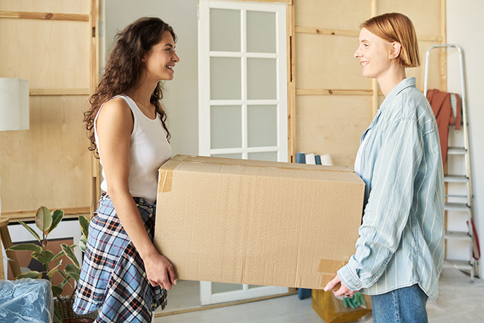 Two women smiling and carrying a large cardboard box together, moving out of an apartment. Two women smiling and carrying a large cardboard box together, moving out of an apartment.
