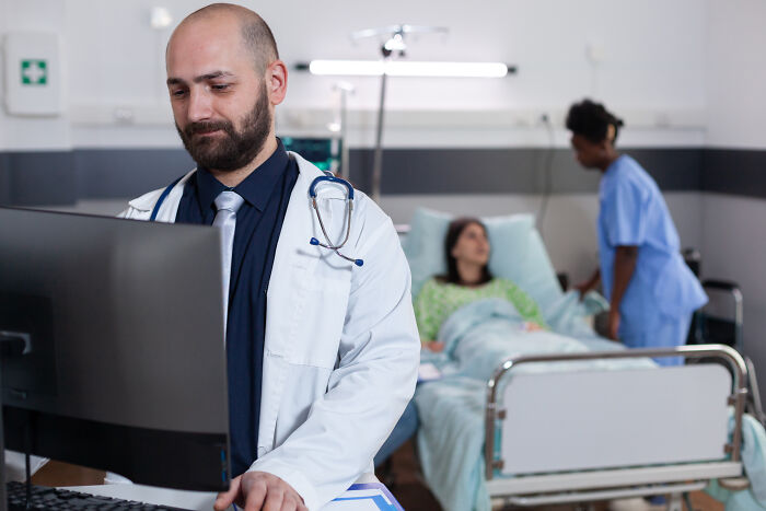 A doctor at a computer, with a nurse and patient in the background, handling pain reactions.
