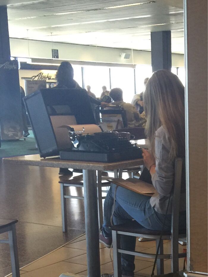 A woman typing on a vintage typewriter at an airport, one of the weird things spotted at airports.
