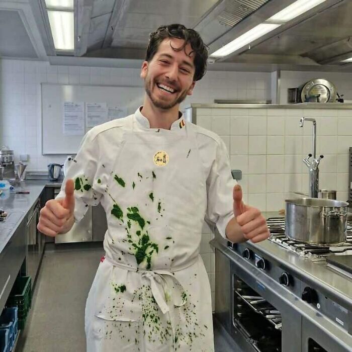 A smiling chef with green food stains on his white uniform, giving two thumbs up for a kitchen fail.