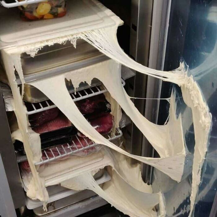 A refrigerator with a severe kitchen fail, showing dough oozing and dripping from multiple shelves.