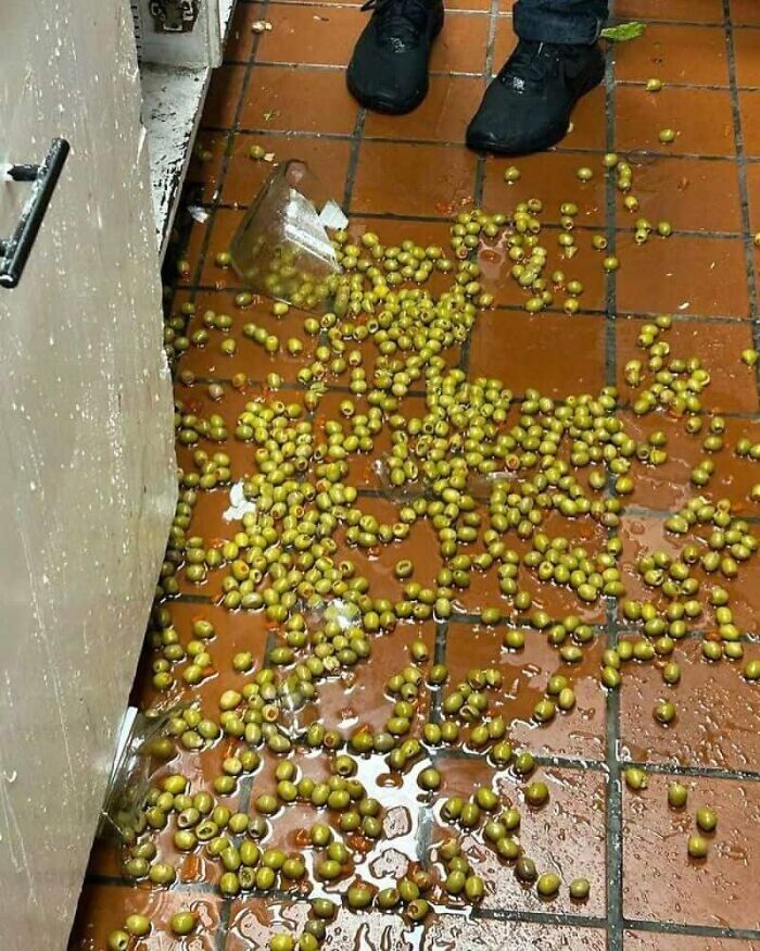 A kitchen fail with countless green olives spilled across a wet, tiled floor, next to a persons feet.